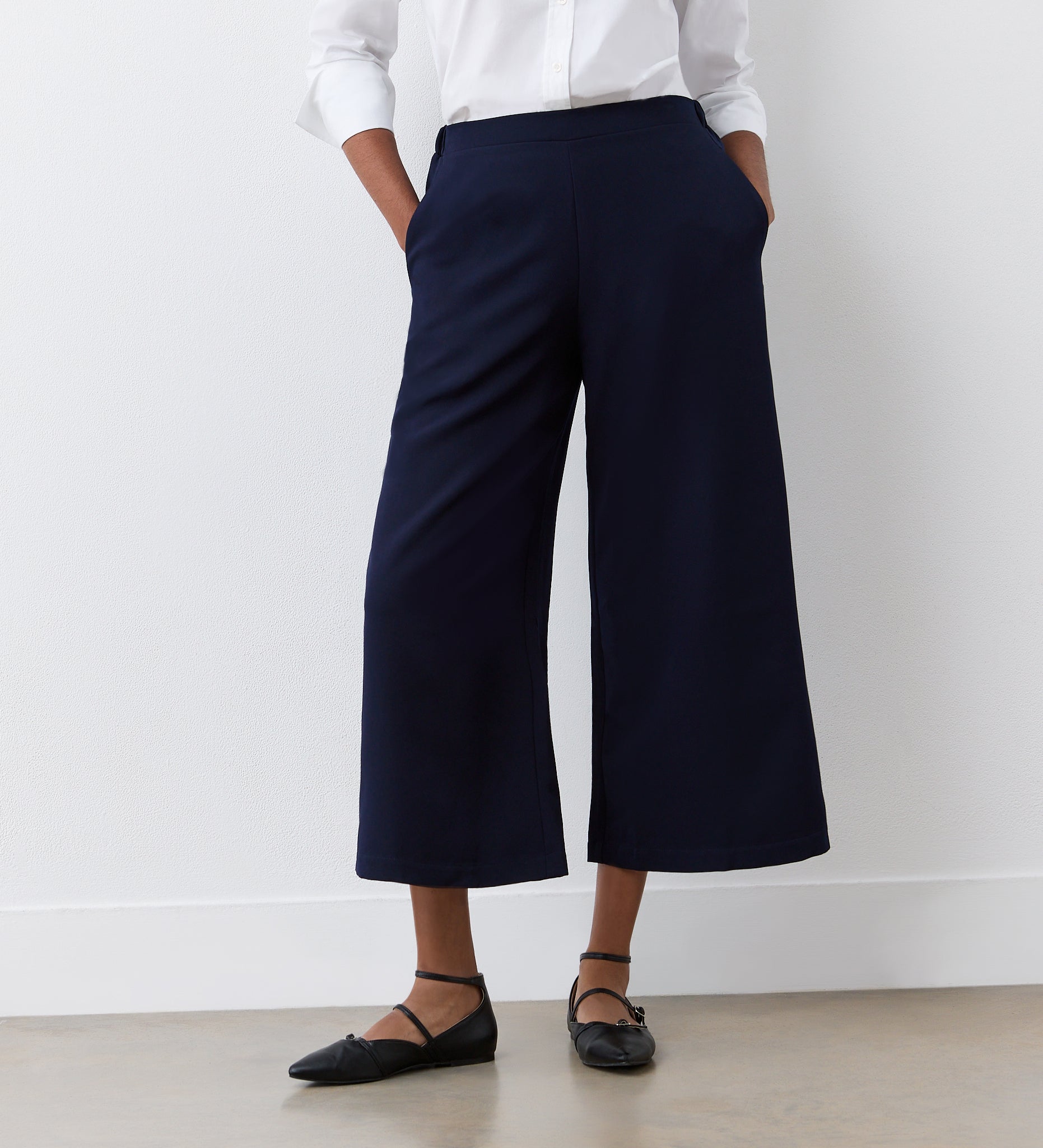 A person stands against a white wall with hands in their pockets, wearing a white long-sleeve blouse, black flats, and the Simone Navy Cropped Wide Leg Trousers featuring an elasticated waistband.
