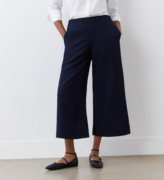 A person stands against a white wall with hands in their pockets, wearing a white long-sleeve blouse, black flats, and the Simone Navy Cropped Wide Leg Trousers featuring an elasticated waistband.