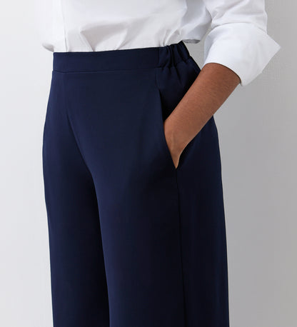 A person stands against a light wall wearing a white long-sleeve shirt and the Simone Navy Cropped Wide Leg Trousers with an elasticated waistband—an effortlessly stylish wardrobe essential for any season.