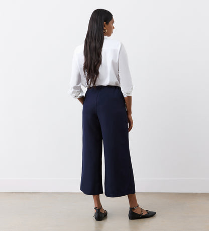 A woman with long dark hair stands facing away, wearing a white long-sleeve blouse and the Simone Navy Cropped Wide Leg Trousers, paired with black pointed-toe ankle strap flats, against a plain white wall and light floor.
