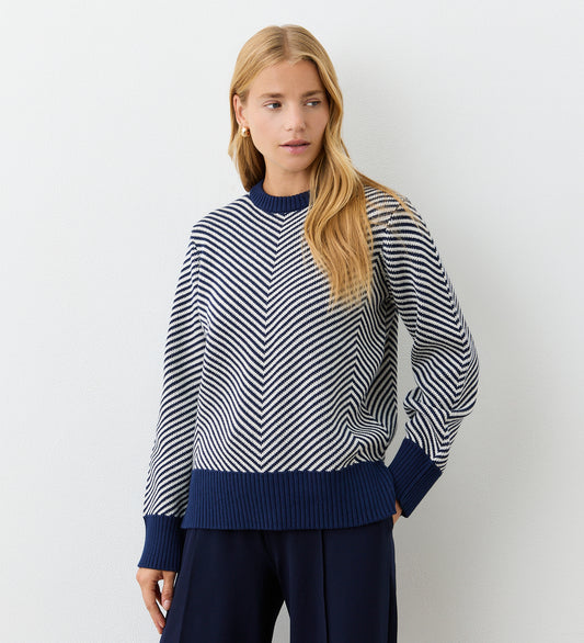 A woman with long blonde hair wears the Stella Navy Chevron Cotton Crew Neck Jumper with white stripes and dark pants, standing against a plain white background and looking to her left.