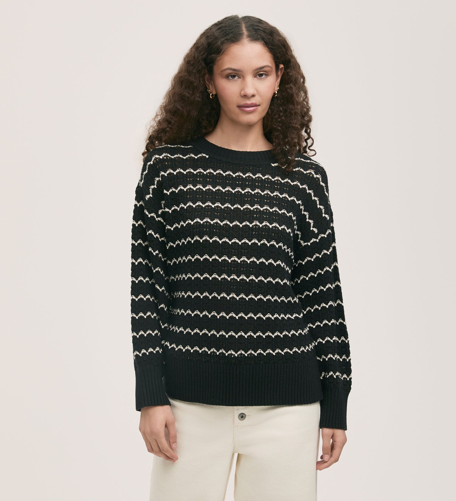 A woman with curly hair wears the Sansa Black Crochet Jumper, a relaxed fit sweater with a white striped zig-zag design, and off-white pants, standing against a neutral background.