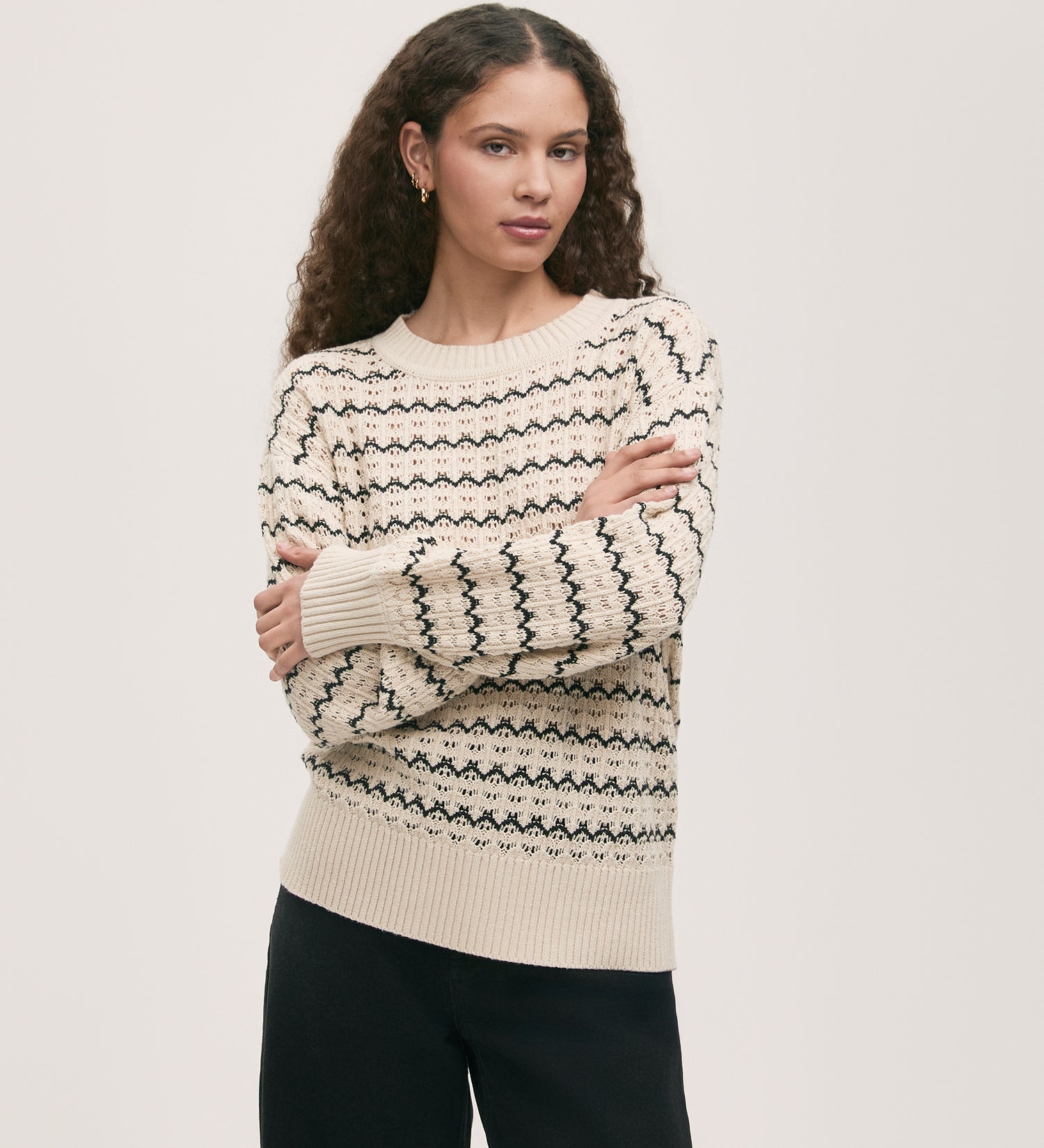 A woman with long curly hair wears the Sansa Ivory Crochet Jumper, featuring ribbed detailing and black geometric patterns, paired with black pants. She stands against a plain light background with her arms crossed.