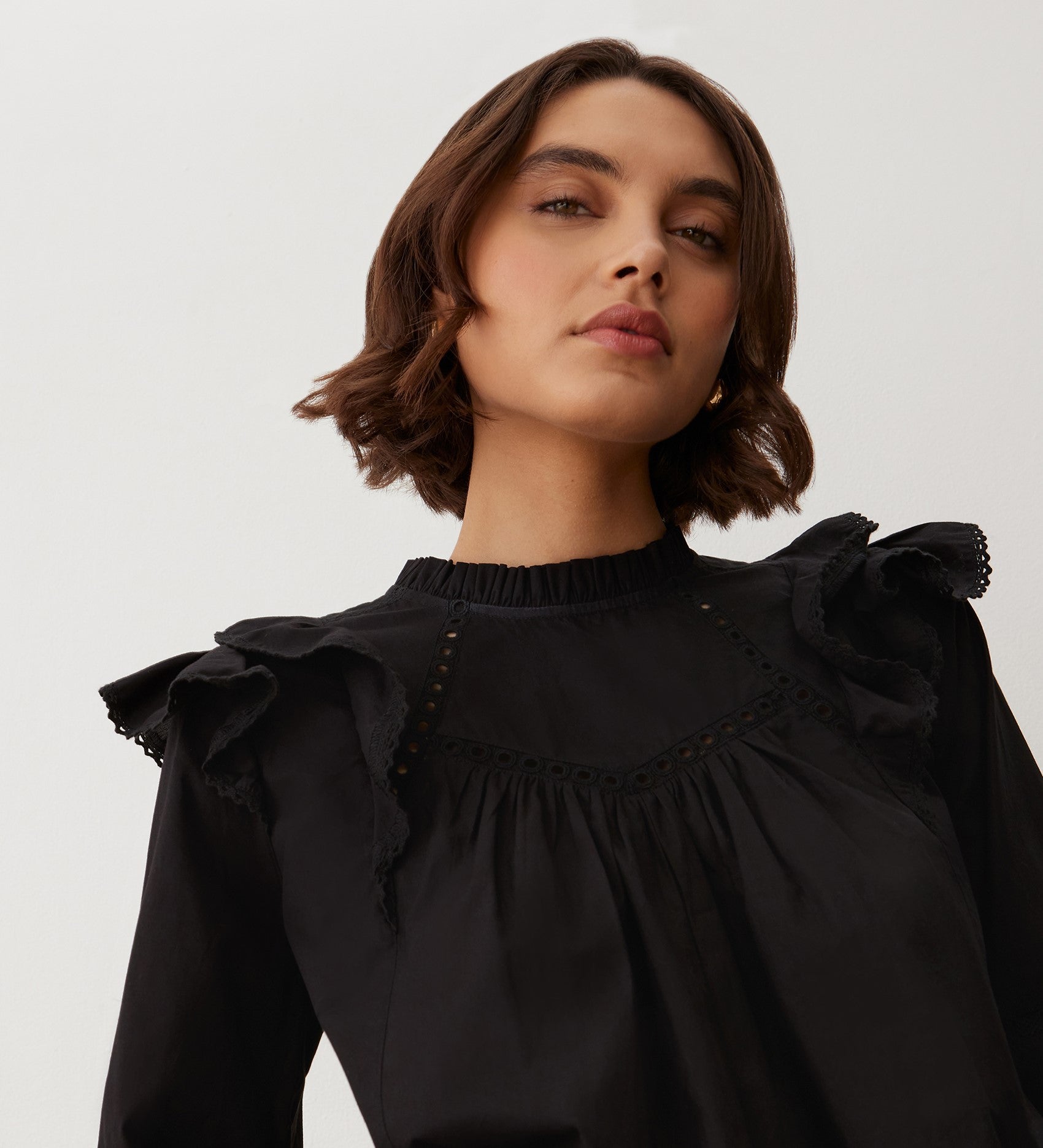A woman with short brown hair confidently models the Zelda Black Cotton Blouse, featuring blouson sleeves and frill edge lace details, against a plain light background.