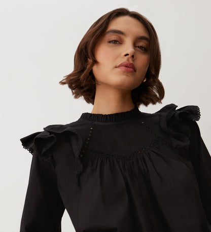 A woman with short brown hair confidently models the Zelda Black Cotton Blouse, featuring blouson sleeves and frill edge lace details, against a plain light background.