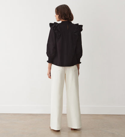 A woman faces away indoors, wearing the Zelda Black Cotton Blouse with blouson sleeves and ruffled shoulders, paired with white wide-leg pants. She stands against a plain white wall and light-colored floor.