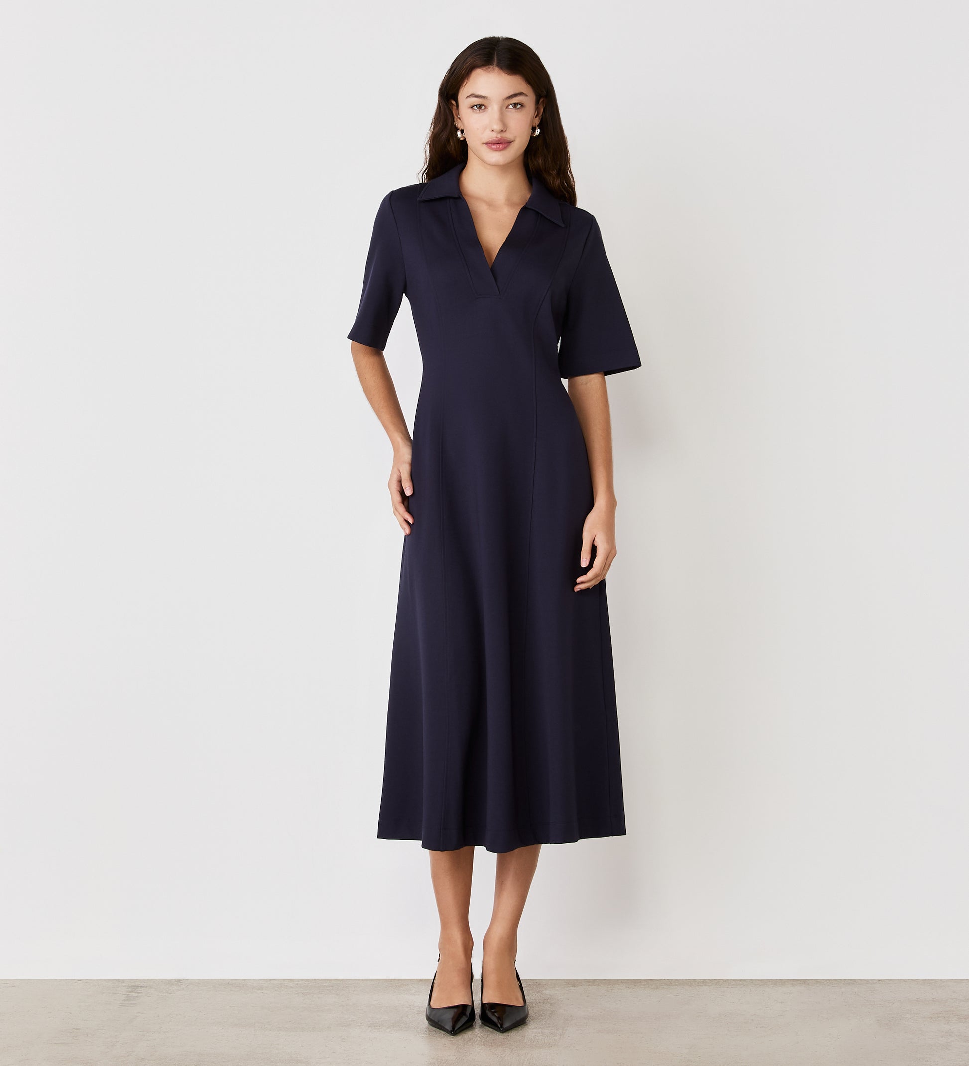A woman with long dark hair and a neutral expression stands against a white wall, wearing the Trinity Navy Ponte Jersey Midi Dress with short sleeves, a collar, and black pointed-toe heels.