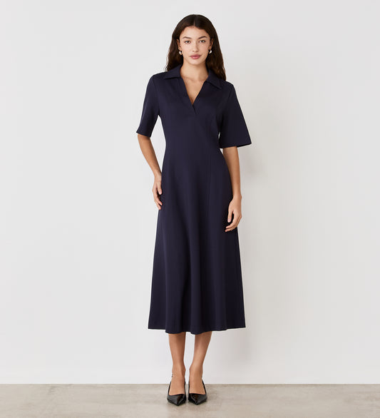 A woman with long dark hair and a neutral expression stands against a white wall, wearing the Trinity Navy Ponte Jersey Midi Dress with short sleeves, a collar, and black pointed-toe heels.