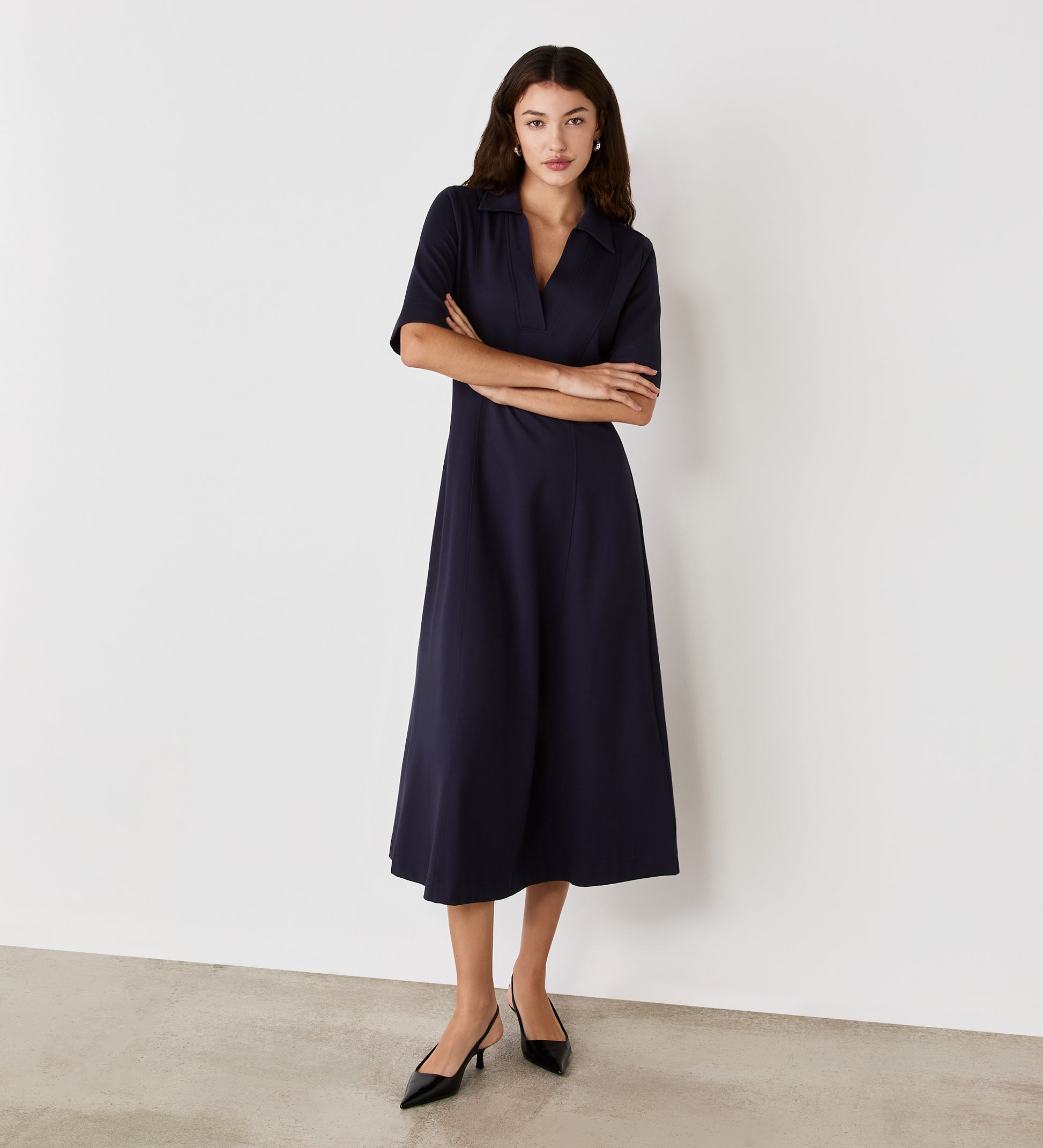 A woman stands against a white wall, wearing the Trinity Navy Ponte Jersey Midi Dress with short sleeves and black pointed-toe heels. Her arms are crossed as she looks at the camera with a neutral expression.