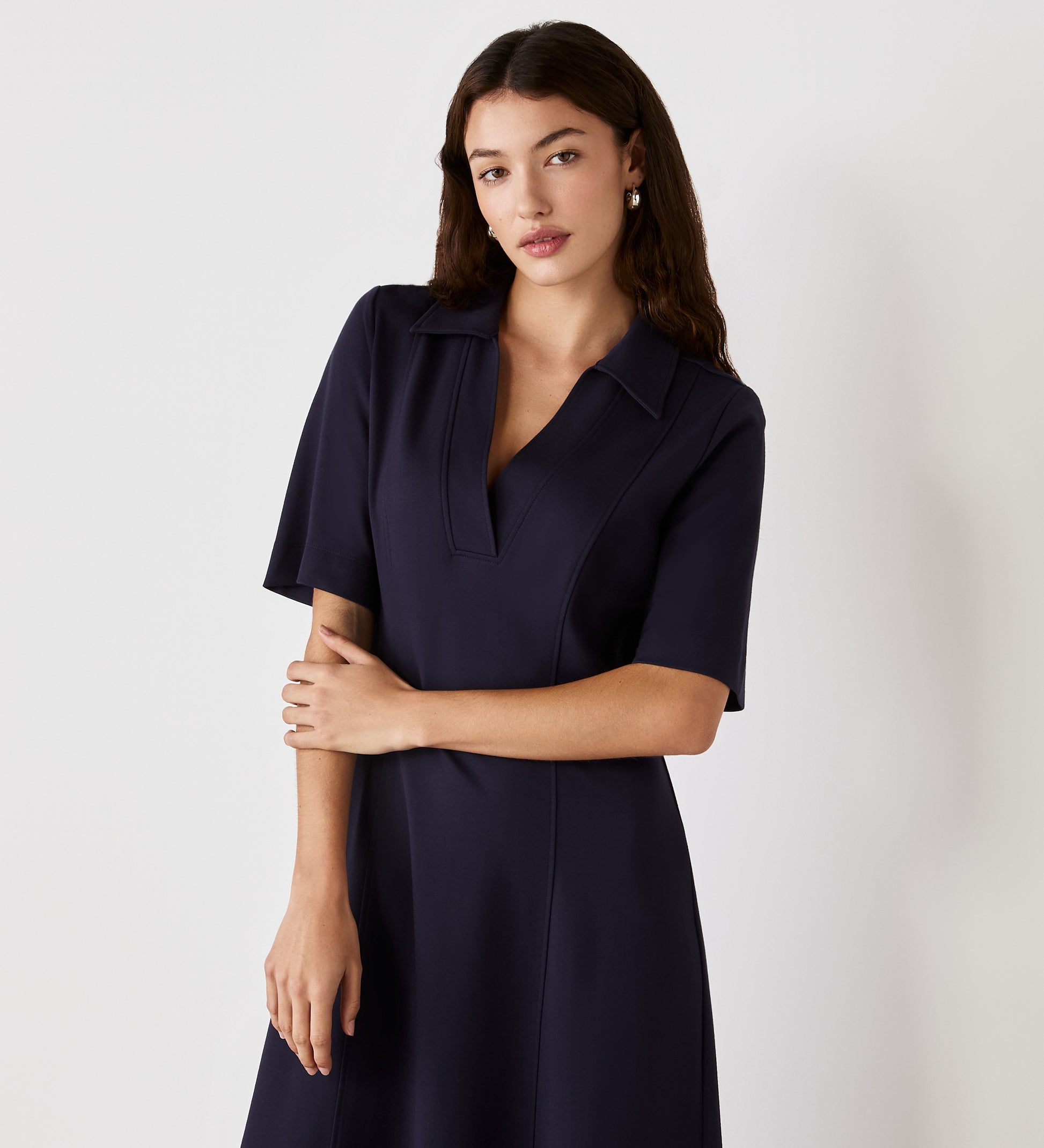 A woman with long brown hair wears the Trinity Navy Ponte Jersey Midi Dress with short sleeves, standing against a plain light background. She has one arm crossed and maintains a neutral expression.