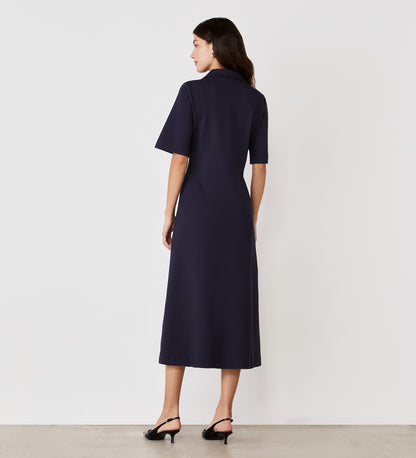 A woman stands facing away, wearing the Trinity Navy Ponte Jersey Midi Dress with short sleeves and black heeled sandals, against a plain white background.