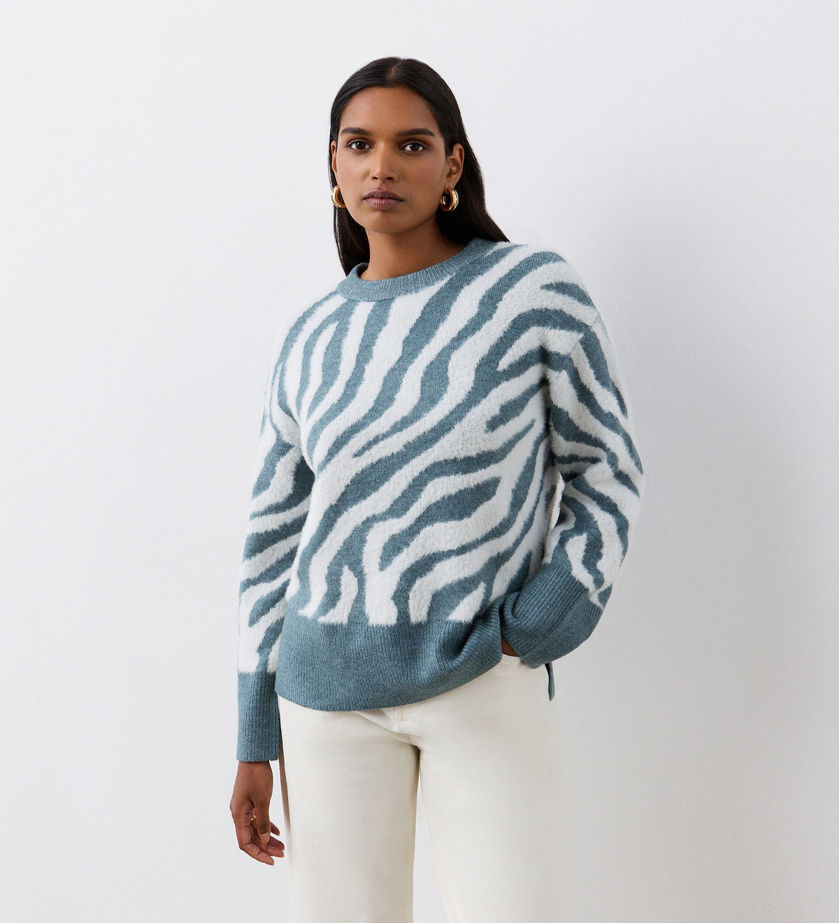 A woman with long dark hair and hoop earrings stands against a white background, wearing the Tully Duck Egg Zebra Knitted Jumper with light-colored pants. She looks directly at the camera with a neutral expression.