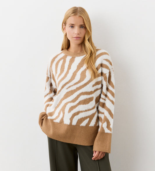 A woman with long blonde hair wears the Tully Tan Zebra Knitted Jumper, featuring a beige and white animal print with brown trim, paired with dark pants, standing against a plain white background.