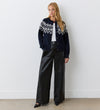 A woman stands against a white wall, wearing the Violet Navy Cotton Cardigan with a white geometric pattern and round neckline, paired with a white top, wide-leg black leather pants, and black pointed-toe shoes.