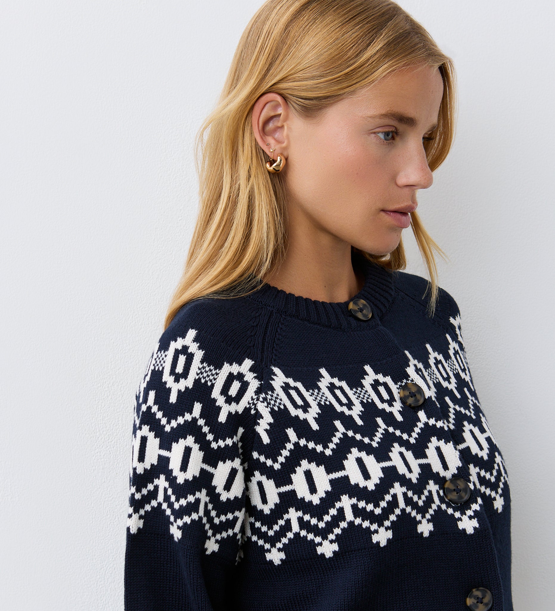 A woman with blonde hair and gold hoop earrings wears the Violet Navy Cotton Cardigan, featuring a round neckline and brown buttons, standing against a plain white wall.