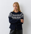 A woman with long blonde hair stands against a white wall, arms crossed, wearing the Violet Navy Cotton Cardigan featuring a white geometric fair isle pattern and round neckline, looking at the camera with a neutral expression.