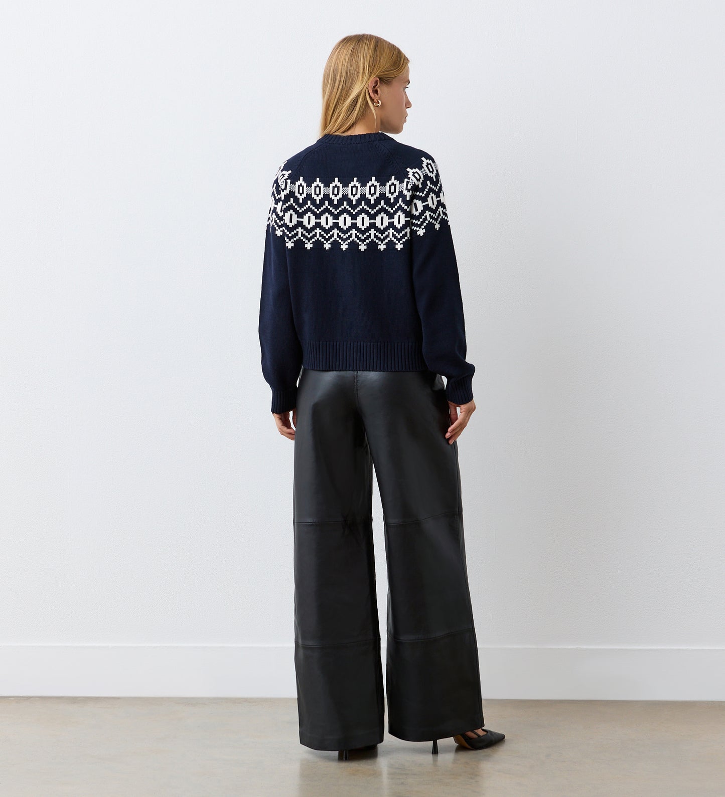 A woman with blonde hair faces a white wall, wearing the Violet Navy Cotton Cardigan with a round neckline and wide-leg black leather pants paired with black heels.