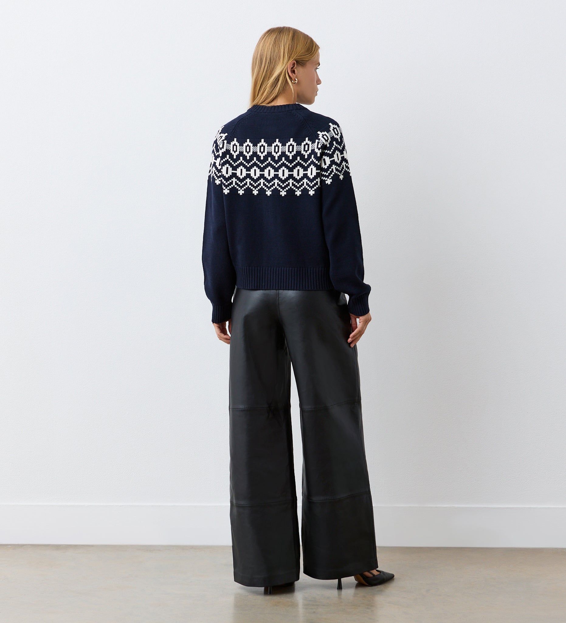 A woman with blonde hair faces a white wall, wearing the Violet Navy Cotton Cardigan with a round neckline and wide-leg black leather pants paired with black heels.
