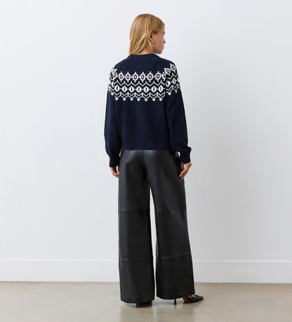 A woman with blonde hair faces a white wall, wearing the Violet Navy Cotton Cardigan with a round neckline and wide-leg black leather pants paired with black heels.