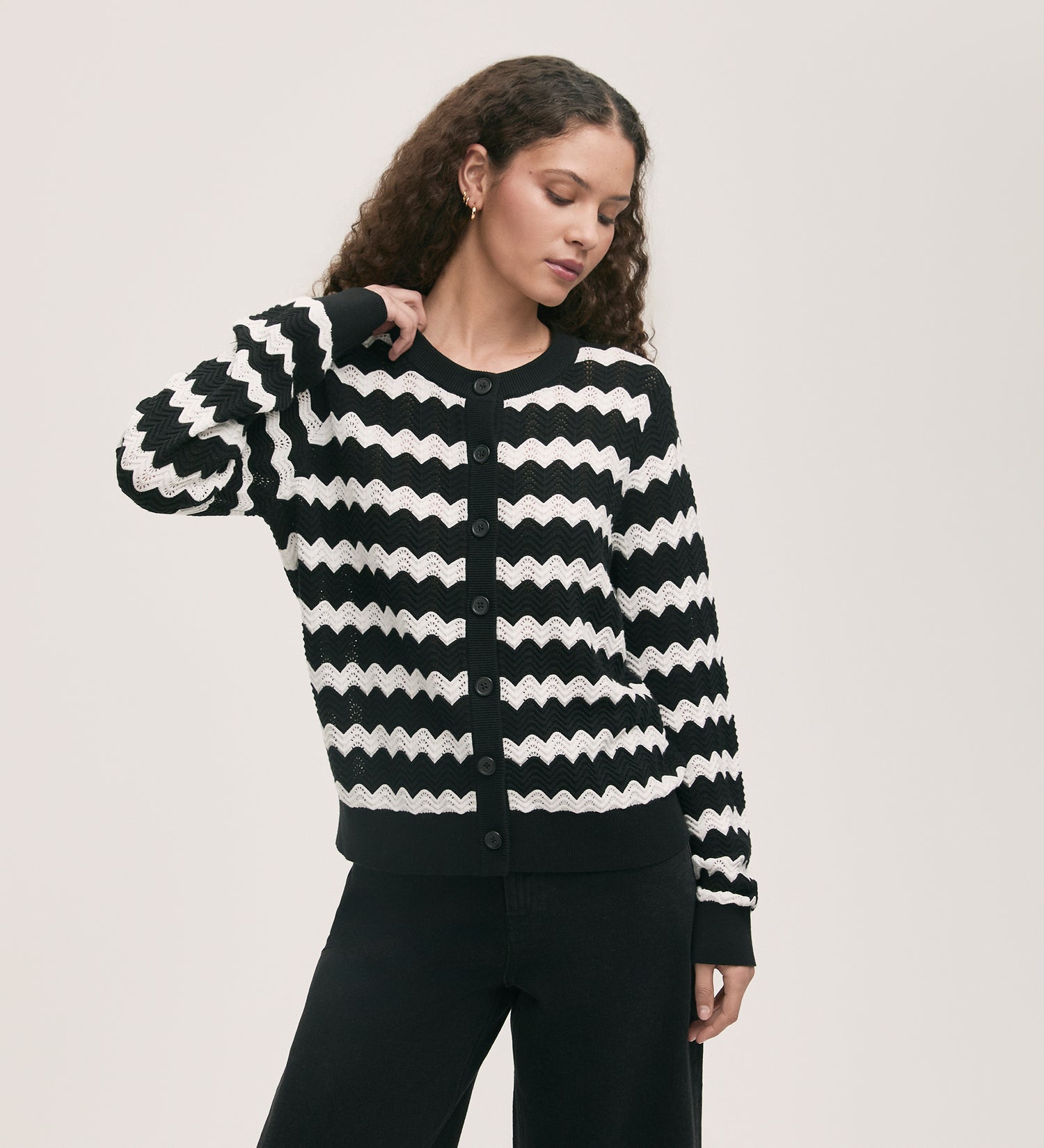 A woman with long curly hair wears the Velma Black Stripe Crochet Cardigan, featuring a black and white zigzag pattern and button-up front, paired with black pants. She stands against a plain light background, looking down with one hand on her head.
