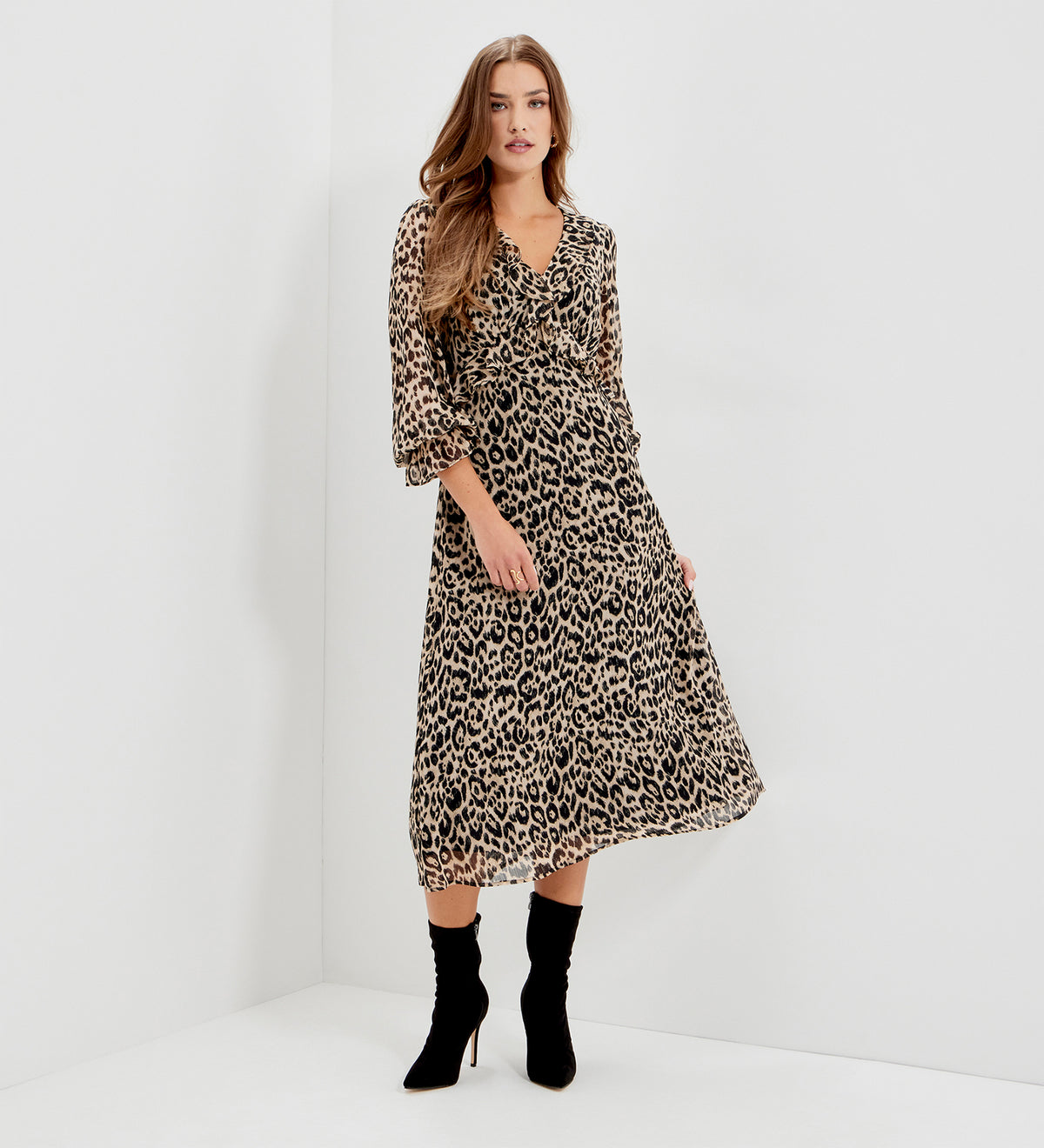 A woman modeling Veronica Natural Animal Ruffle Empire Waist Midi Dress, paired with black ankle boots, standing in a white studio corner.