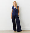 A woman stands against a light wall wearing the Winnie Navy Sleeveless Cardigan over a white shirt, paired with wide-leg navy pants and cream-colored shoes.