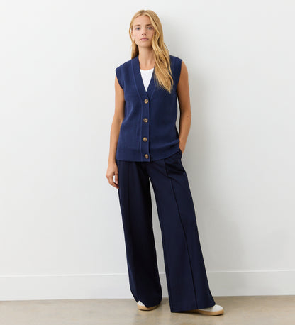 A woman stands against a light wall wearing the Winnie Navy Sleeveless Cardigan over a white shirt, paired with wide-leg navy pants and cream-colored shoes.