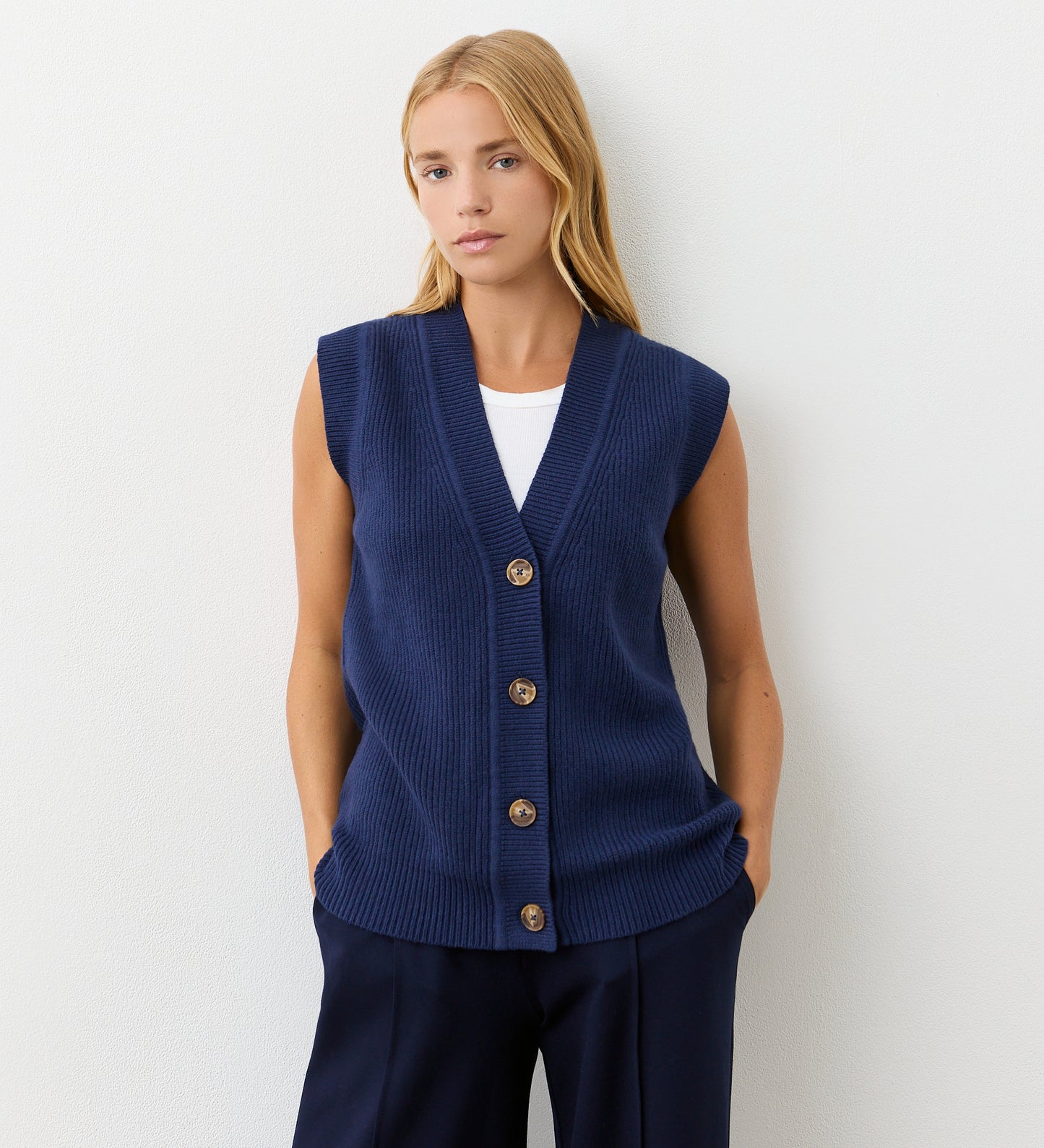 A woman with long blonde hair wears the Winnie Navy Sleeveless Cardigan with a v-neckline and button front over a white shirt and navy pants, standing against a plain white background with her hands in her pockets.