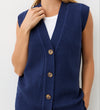 A person wears the Winnie Navy Sleeveless Cardigan—a v-neck, button-front knitted vest—over a white top, standing against a plain white background with one hand touching the collar.