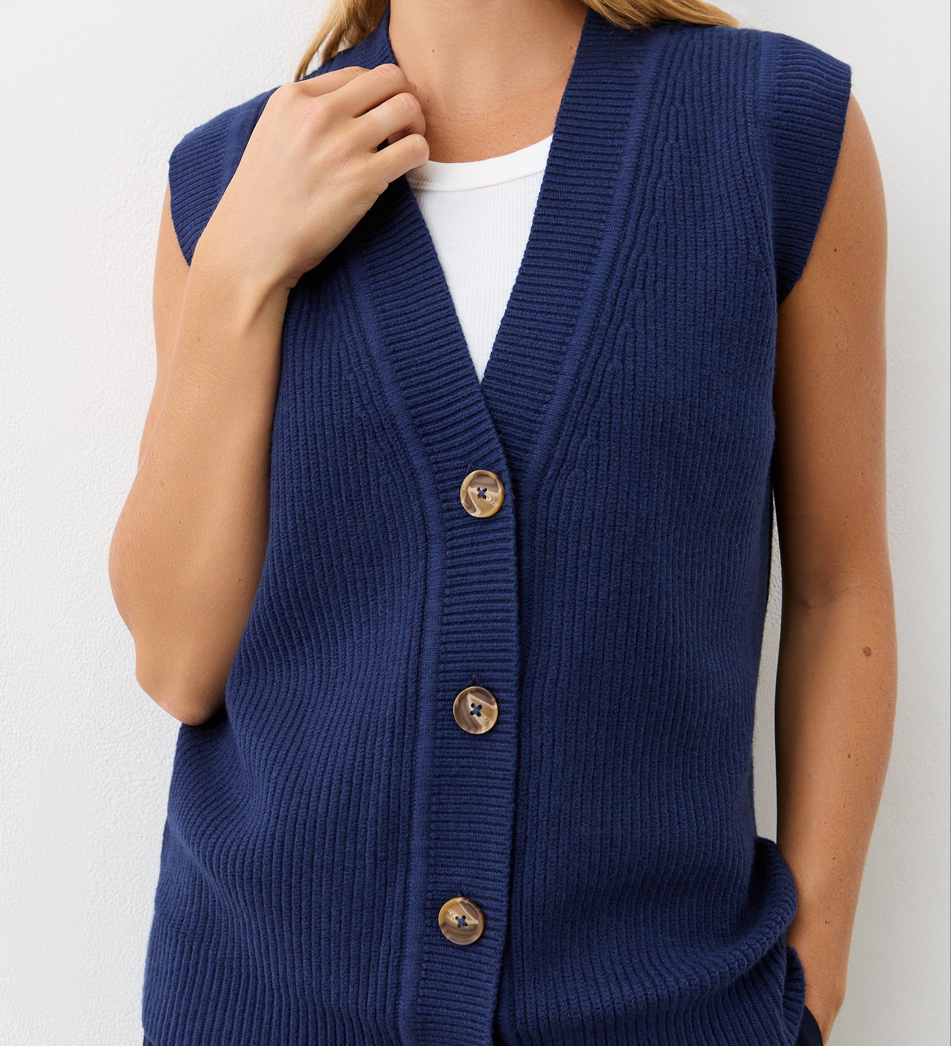 A person wears the Winnie Navy Sleeveless Cardigan—a v-neck, button-front knitted vest—over a white top, standing against a plain white background with one hand touching the collar.