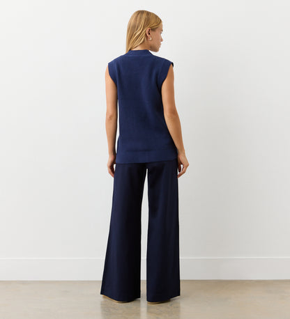 A woman with straight, shoulder-length hair faces a white wall, wearing the Winnie Navy Sleeveless Cardigan and wide-leg navy pants in a minimalistic setting with a light-colored floor.