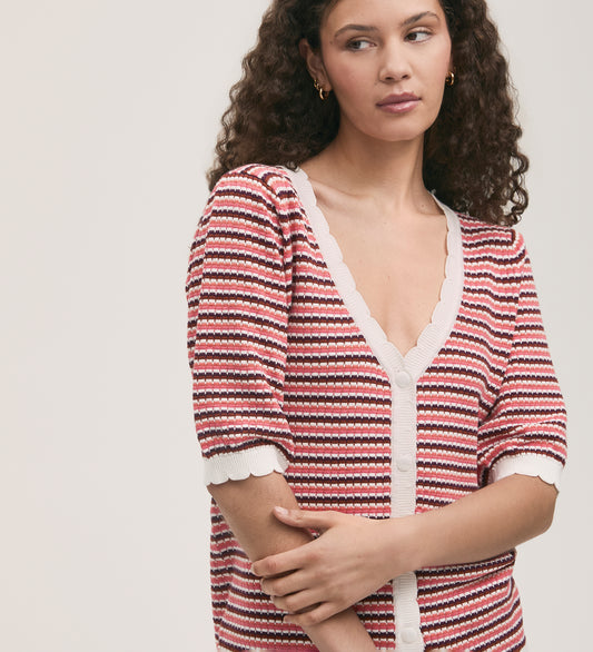 A woman with curly brown hair wears the Willa Pink Stripe Cardigan, a relaxed fit design with scallop ribbed detailing and white buttons. She gazes to the side with a neutral expression against a plain background.