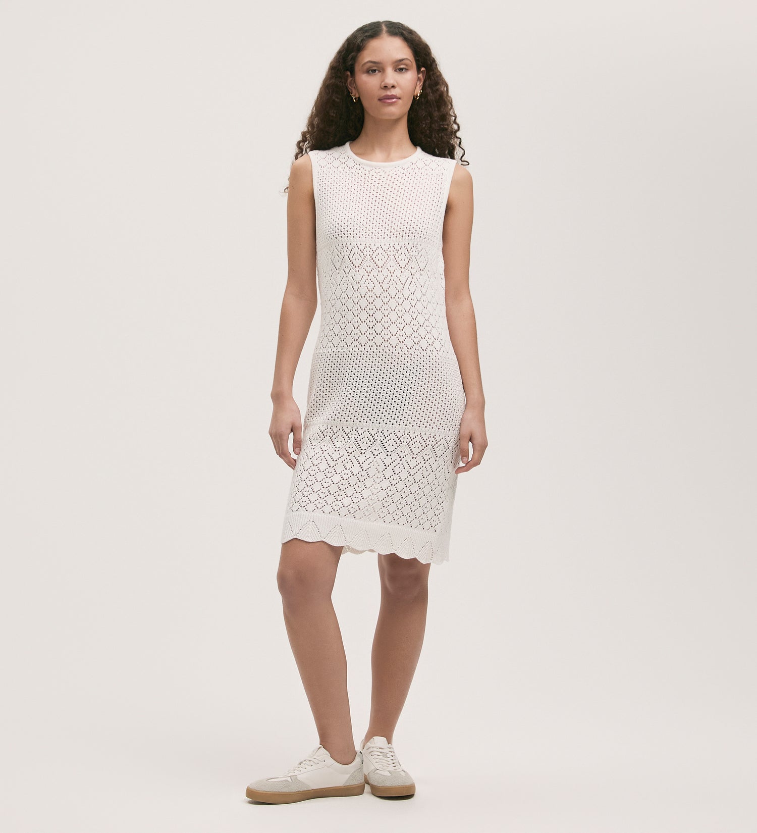 A woman with curly hair models the Zendaya Ivory Crochet Knee Length Dress, showcasing its scallop-detailed hemline. She styles the sleeveless dress with white sneakers while standing against a light background.
