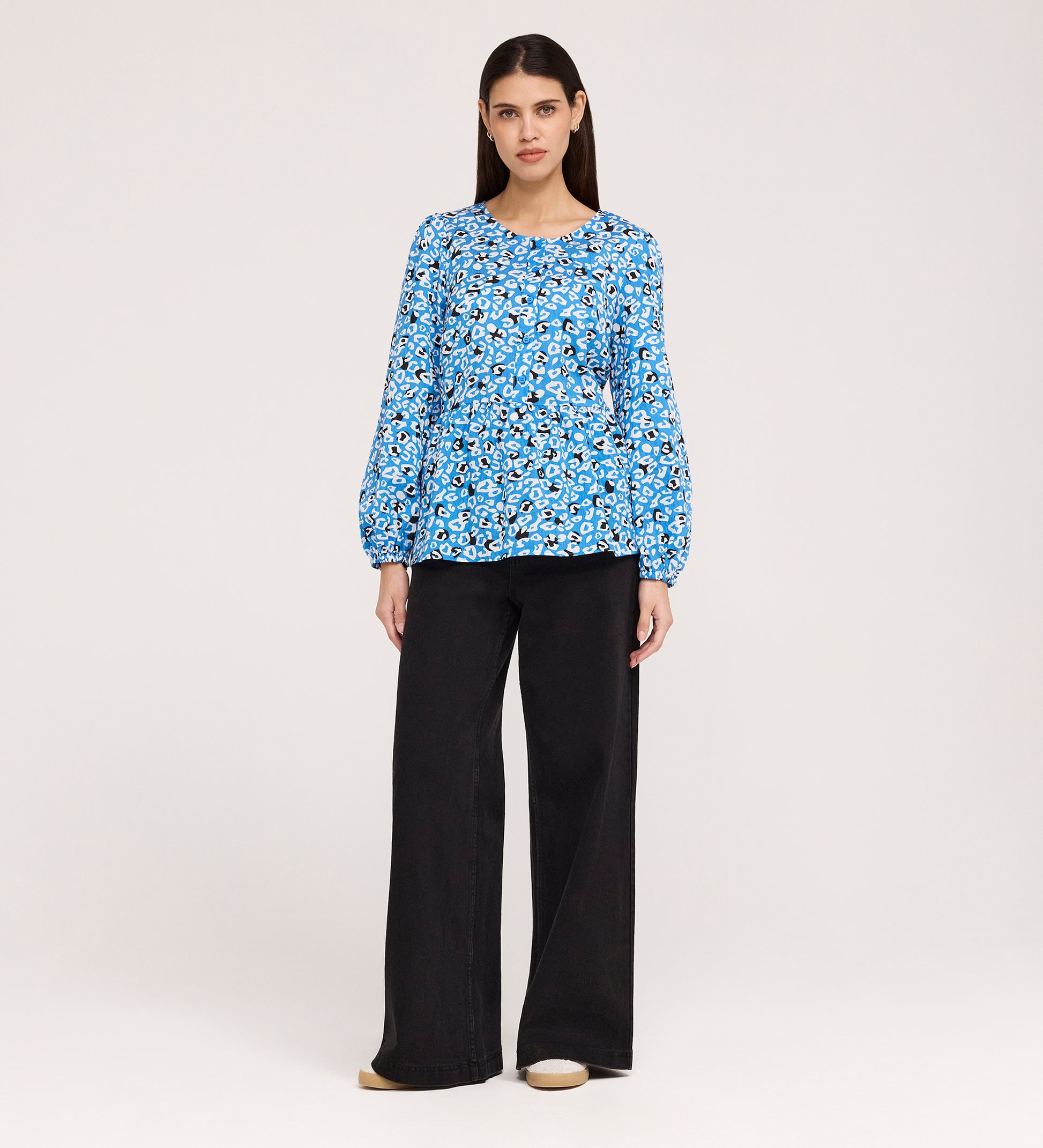 A woman with straight, dark hair stands against a plain background, wearing the Bel Blue Leopard Cotton Blouse with long sleeves and wide-leg black pants, paired with beige shoes. She has a neutral expression.
