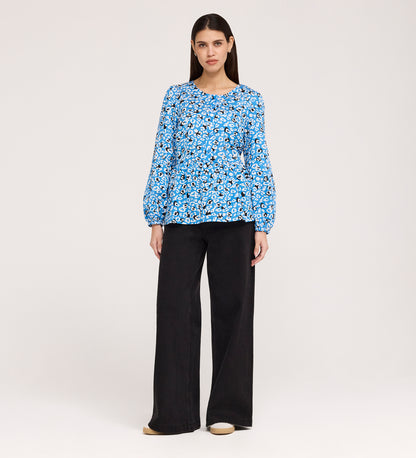 A woman with straight, dark hair stands against a plain background, wearing the Bel Blue Leopard Cotton Blouse with long sleeves and wide-leg black pants, paired with beige shoes. She has a neutral expression.