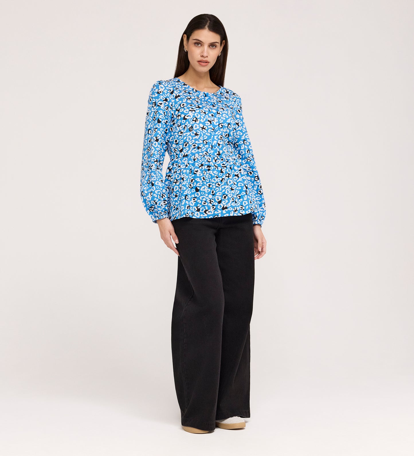 A woman with straight brown hair and a neutral expression stands against a plain background wearing the Bel Blue Leopard Cotton Blouse, featuring long sleeves and a round neck, paired with black wide-leg pants and beige shoes.