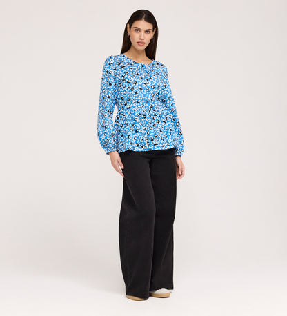 A woman with straight brown hair and a neutral expression stands against a plain background wearing the Bel Blue Leopard Cotton Blouse, featuring long sleeves and a round neck, paired with black wide-leg pants and beige shoes.