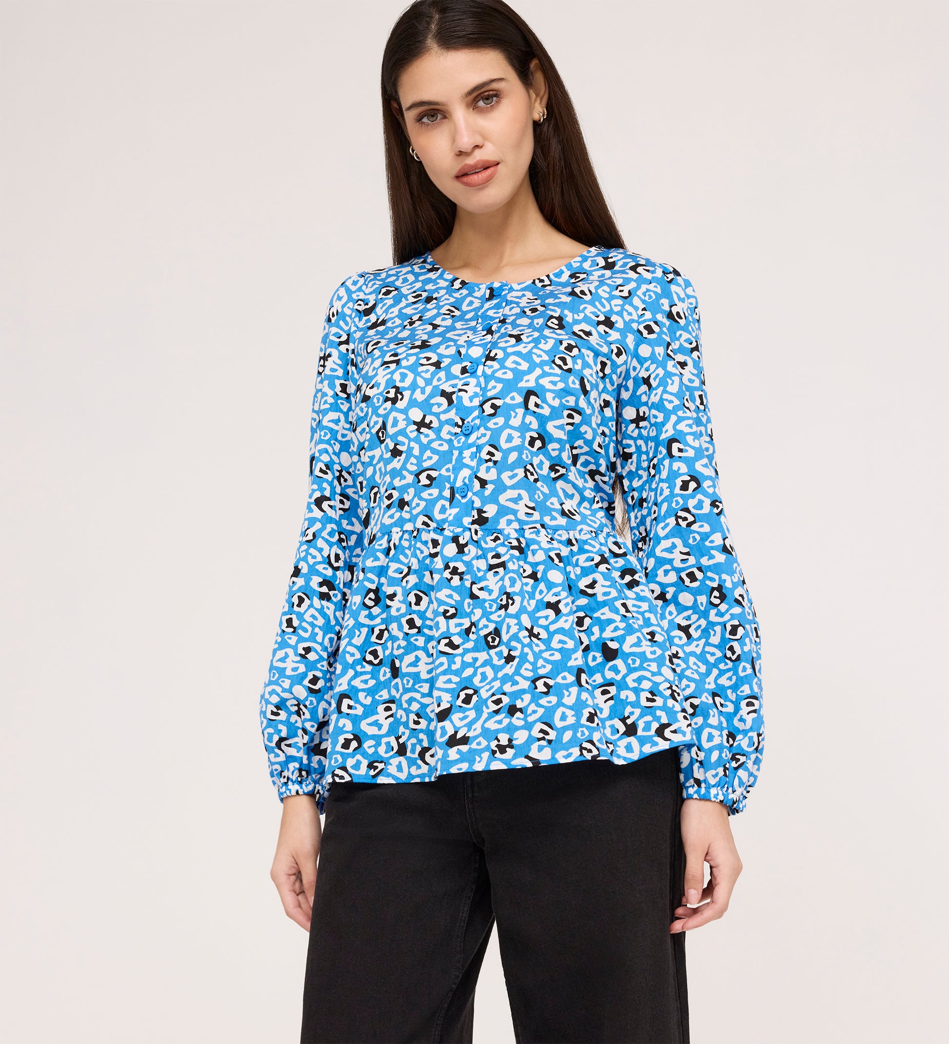 A woman with long dark hair wears the Bel Blue Leopard Cotton Blouse, featuring a round neck, blue, white, and black leopard print, and long sleeves. She pairs it with black pants while standing against a plain light background.