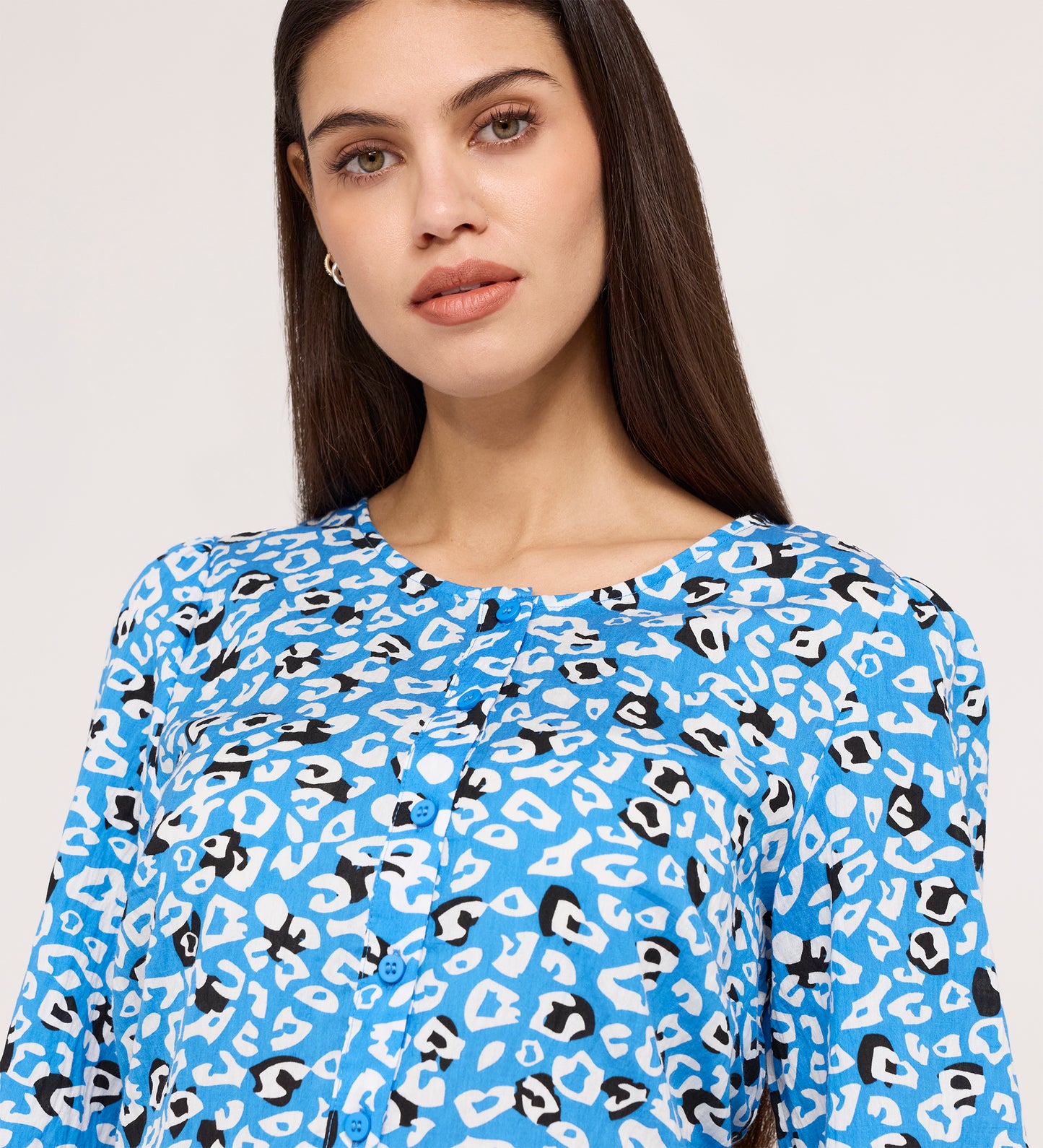 A woman with long brown hair wears the Bel Blue Leopard Cotton Blouse, a blue long-sleeve button-up featuring a white and black abstract animal print, while facing the camera against a plain light background.
