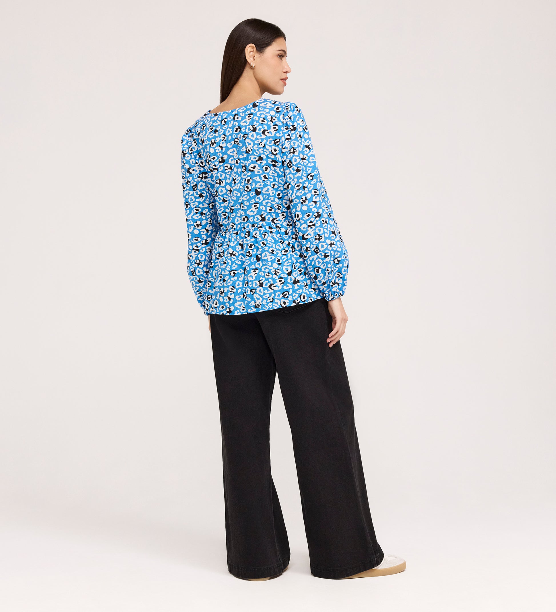 A woman faces away, wearing the Bel Blue Leopard Cotton Blouse with long sleeves, loose black pants, and beige shoes against a plain light background.