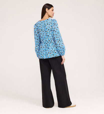 A woman faces away, wearing the Bel Blue Leopard Cotton Blouse with long sleeves, loose black pants, and beige shoes against a plain light background.