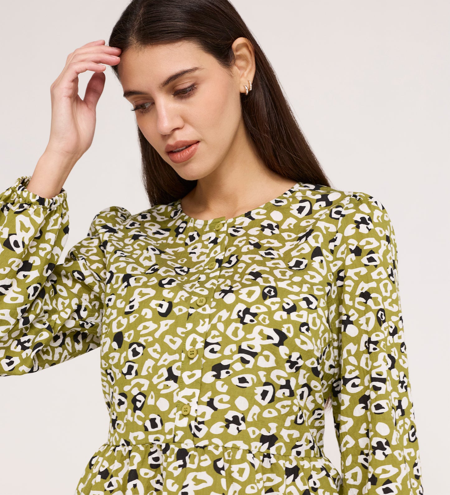 A woman with long brown hair wears the Bel Green Leopard Cotton Blouse, featuring green, white, and black blouson sleeves, as she looks down with one hand raised near her head against a plain background.