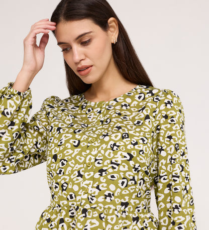 A woman with long brown hair wears the Bel Green Leopard Cotton Blouse, featuring green, white, and black blouson sleeves, as she looks down with one hand raised near her head against a plain background.