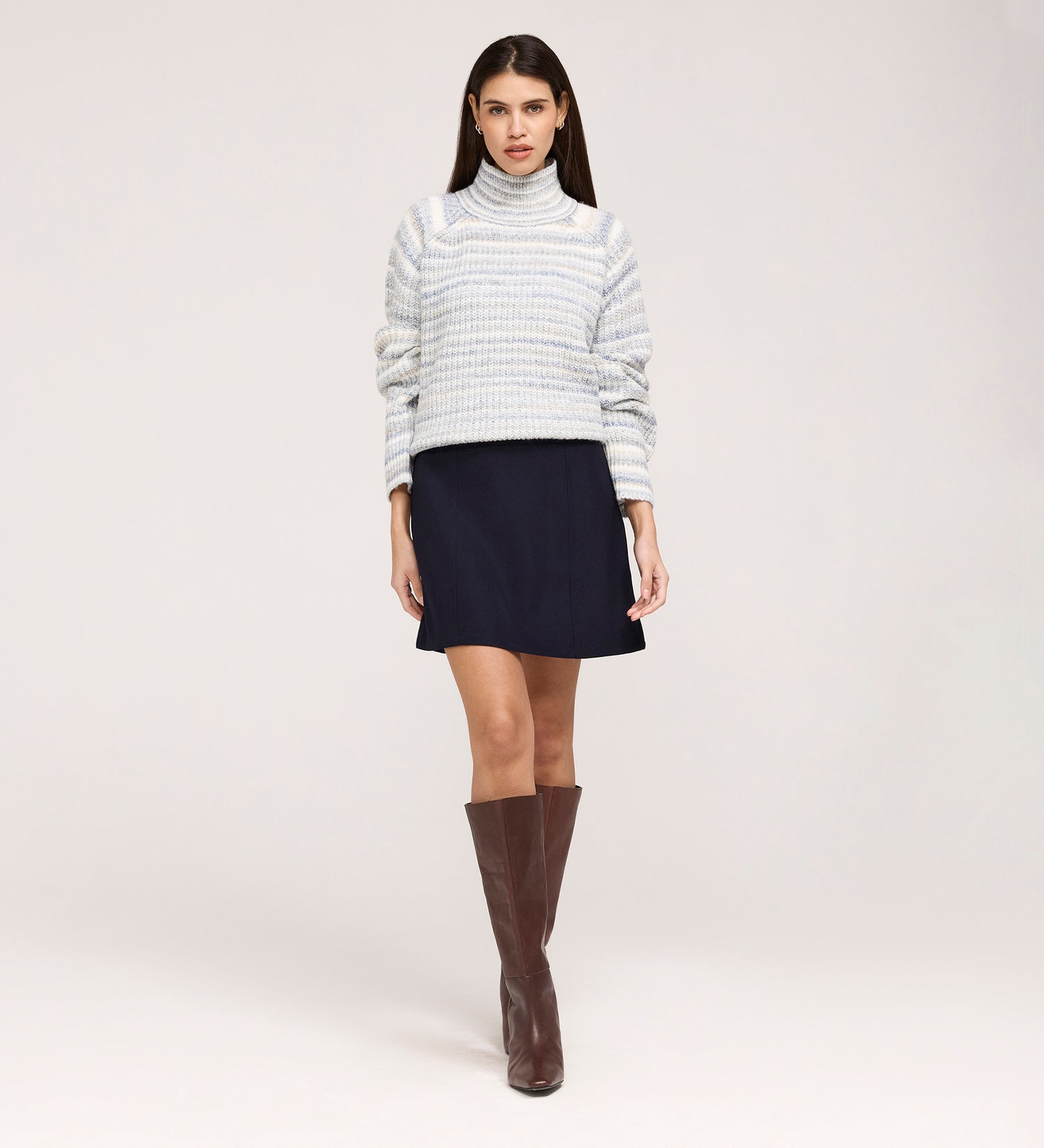 A woman faces forward against a plain light background, wearing a light gray striped turtleneck sweater, knee-high brown boots, and the Kade Navy A-line Gaberdine Twill Mini Skirt.