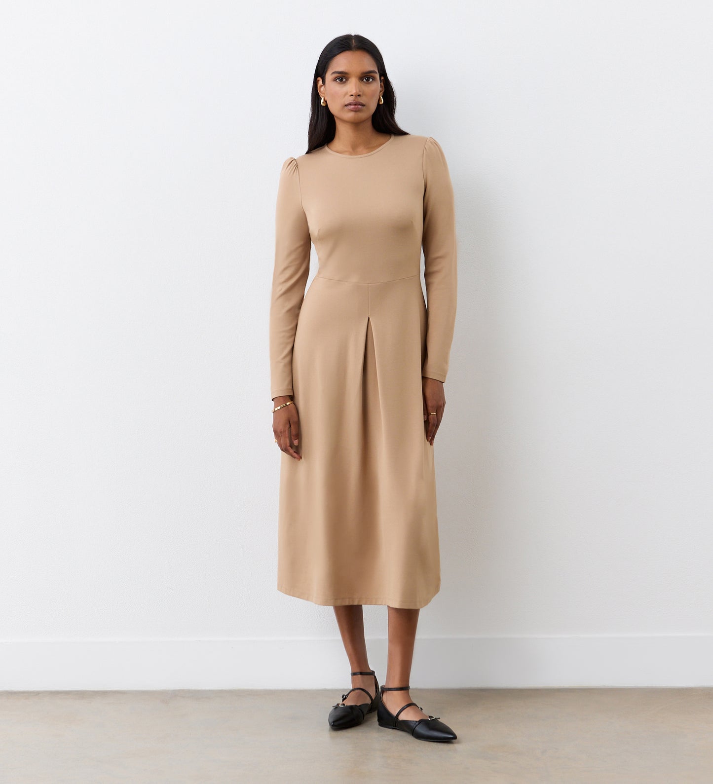 A woman stands against a white wall wearing the Carla Camel Ponte Jersey Midi Dress with long sleeves and a front pleat. Her long, straight hair complements the look, paired with black ankle-strap flats.