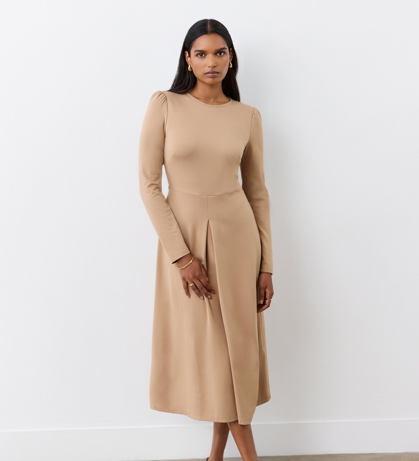 A woman with straight dark hair and minimal jewelry stands against a white wall, wearing the Carla Camel Ponte Jersey Midi Dress featuring a fitted bodice and front pleat detail. She has a neutral expression.