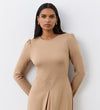 A woman with long dark hair wears the Carla Camel Ponte Jersey Midi Dress, which features subtle puffed shoulders and a front pleat detail. She stands against a white wall, looking at the camera and accessorized with small gold earrings.