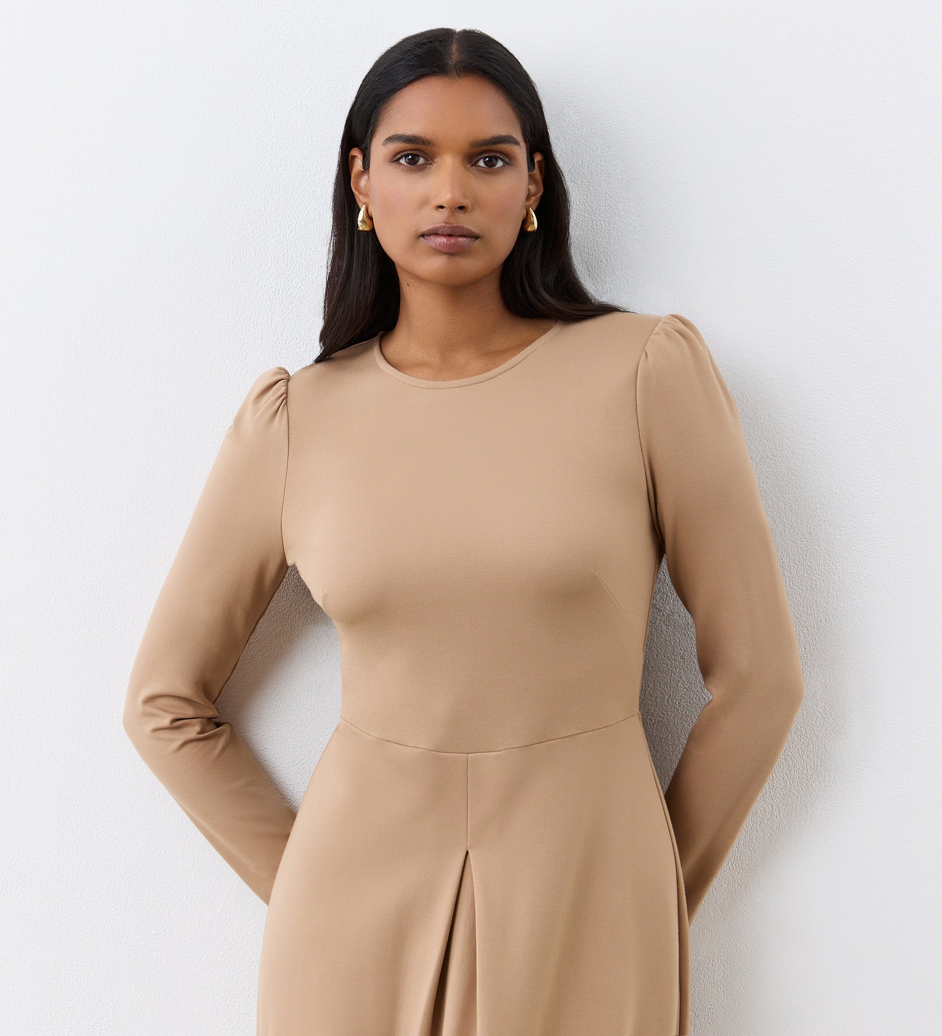 A woman with long dark hair wears the Carla Camel Ponte Jersey Midi Dress, which features subtle puffed shoulders and a front pleat detail. She stands against a white wall, looking at the camera and accessorized with small gold earrings.