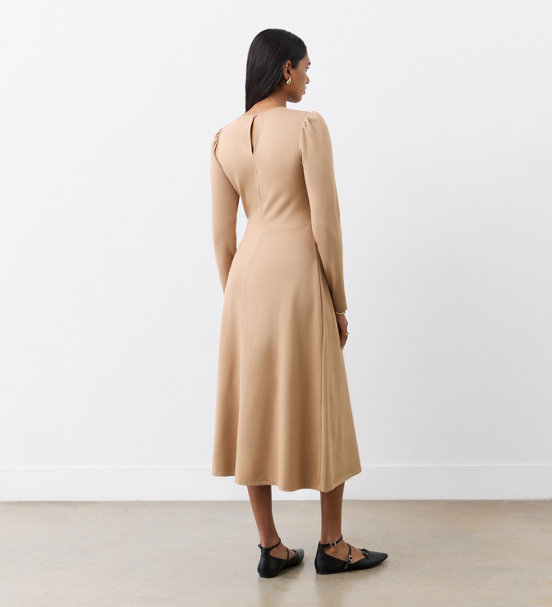 A woman with dark hair, wearing black pointed flats, faces away in the Carla Camel Ponte Jersey Midi Dress featuring a back keyhole. She stands against a plain white wall and light floor.