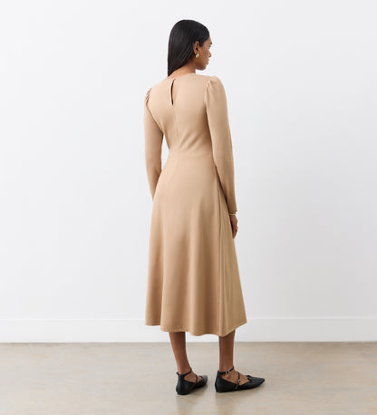 A woman with dark hair, wearing black pointed flats, faces away in the Carla Camel Ponte Jersey Midi Dress featuring a back keyhole. She stands against a plain white wall and light floor.